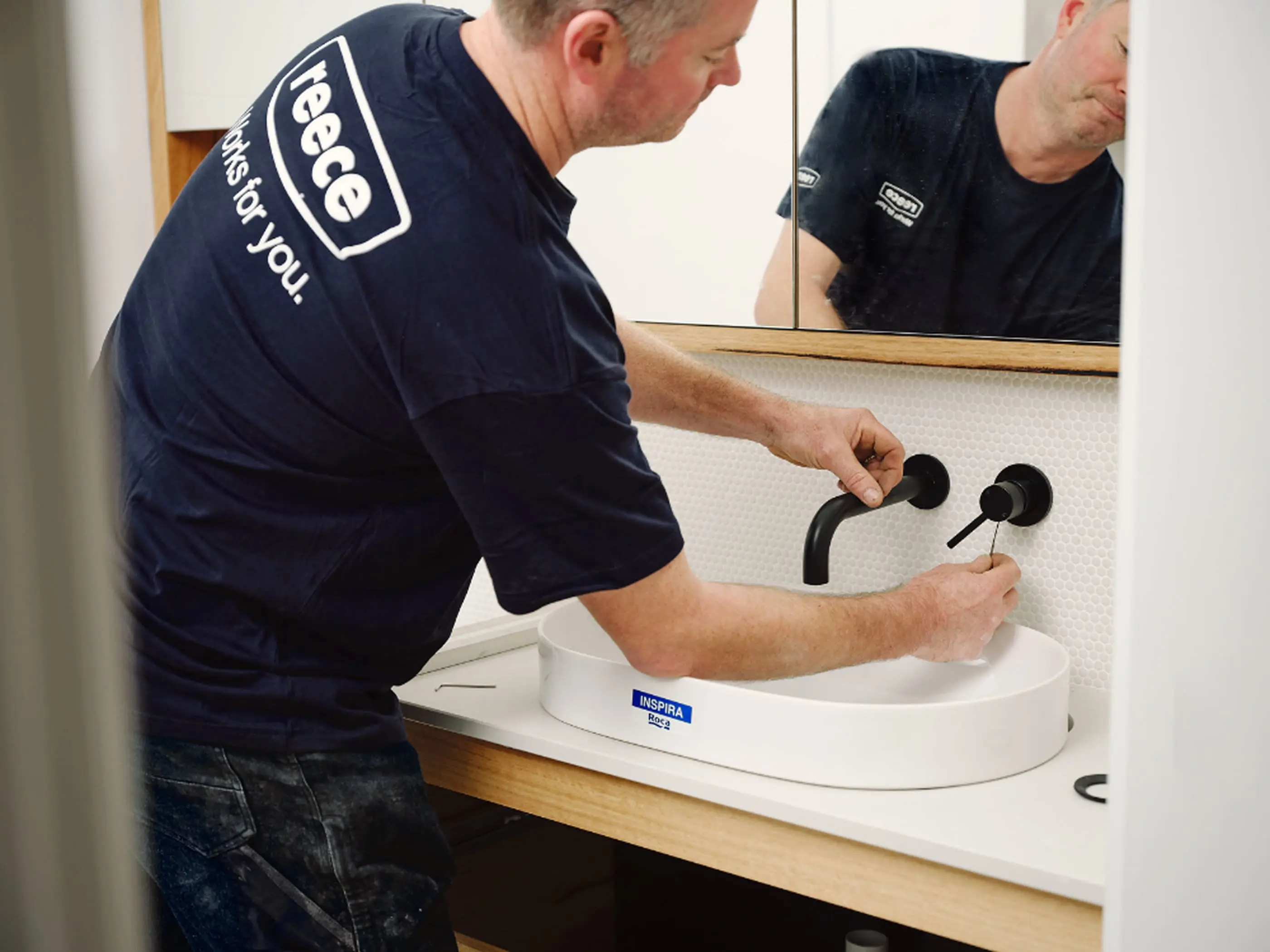 Plumber in navy Reece uniform installing black wall-mounted faucet on white basin in bathroom.