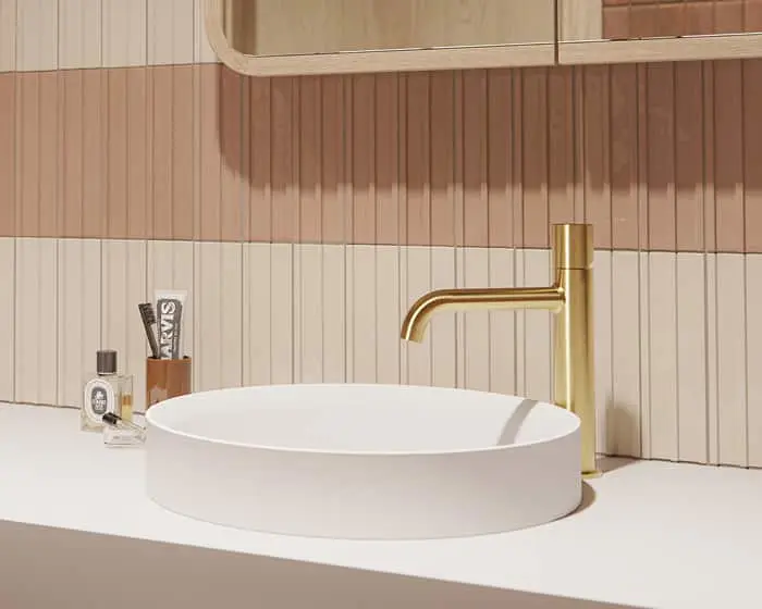 Modern bathroom with white vessel sink, gold faucet, and pink vertical tile backsplash on white countertop.