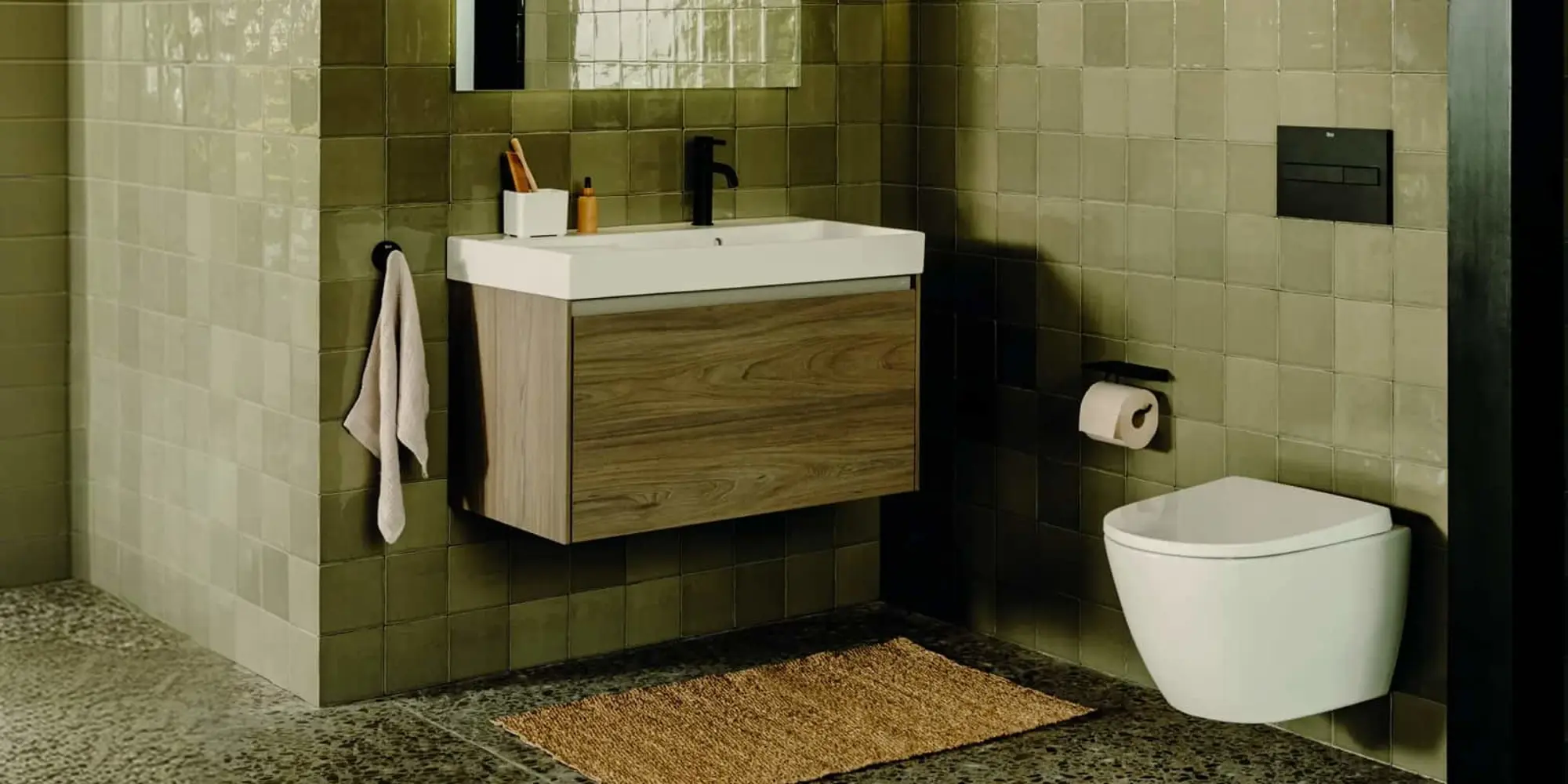 Modern bathroom with green tile walls, wall-mounted sink with wood cabinet, white toilet, and beige floor mat.