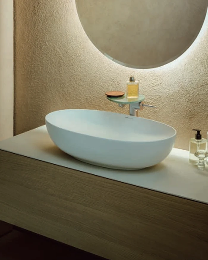 White oval vessel sink on a wood vanity, wall-mounted faucet, round backlit mirror, textured beige wall, soap bottles nearby.