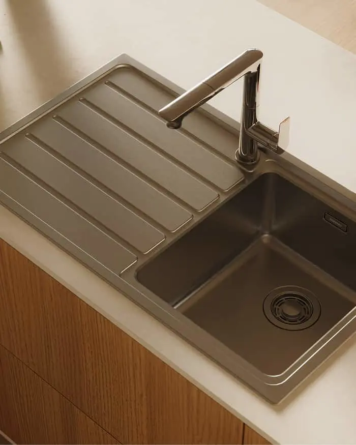 Modern stainless steel kitchen sink with drainboard and sleek faucet installed in white countertop with wooden cabinetry below.