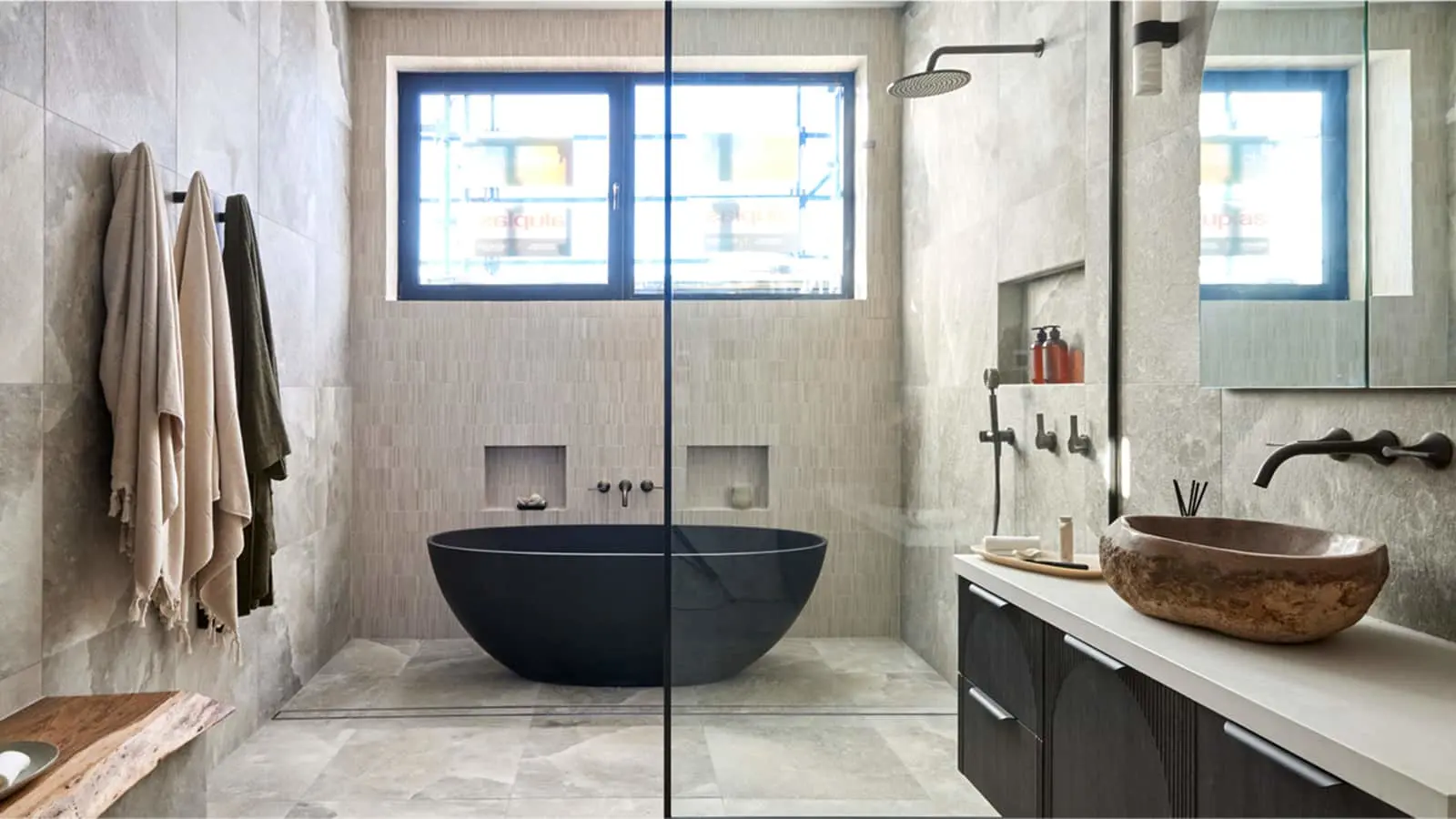 Modern bathroom with black freestanding tub, stone sink, glass shower partition, and light gray tiled walls.