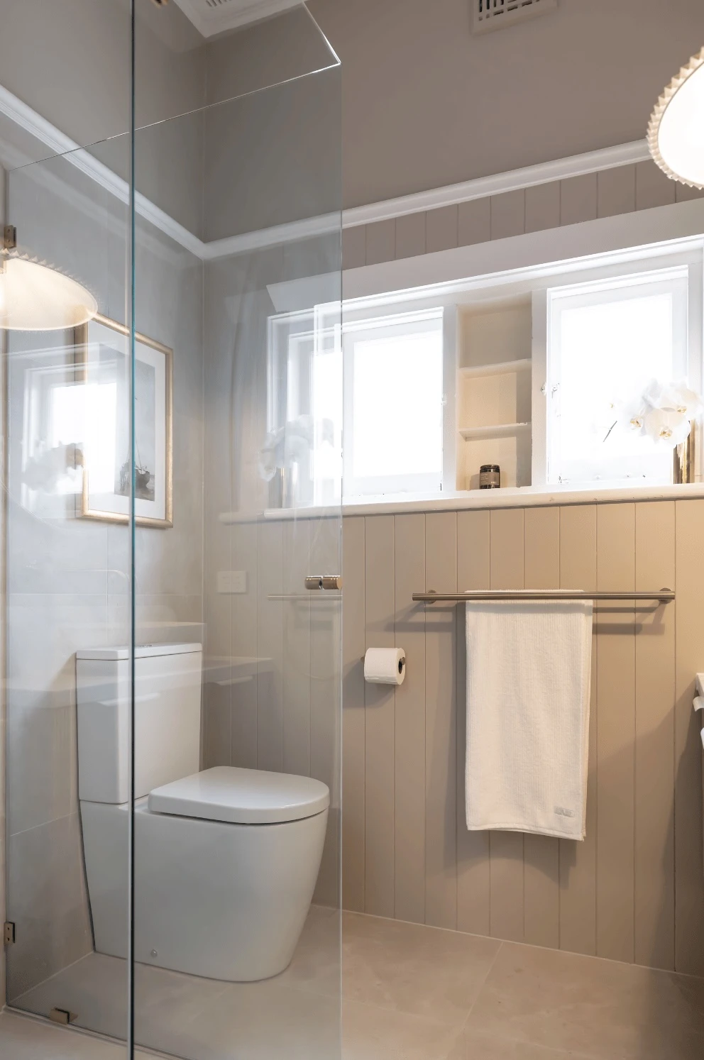 Modern bathroom with glass shower enclosure, white toilet, beige paneled walls, and built-in shelving above towel rail.