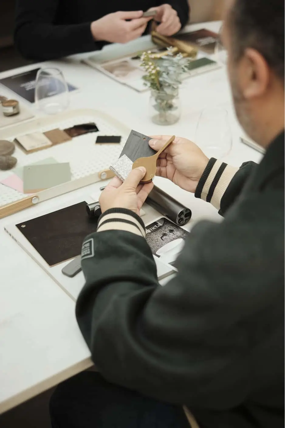 People examining material samples on white table with color swatches, fabric pieces, and a small plant centerpiece.