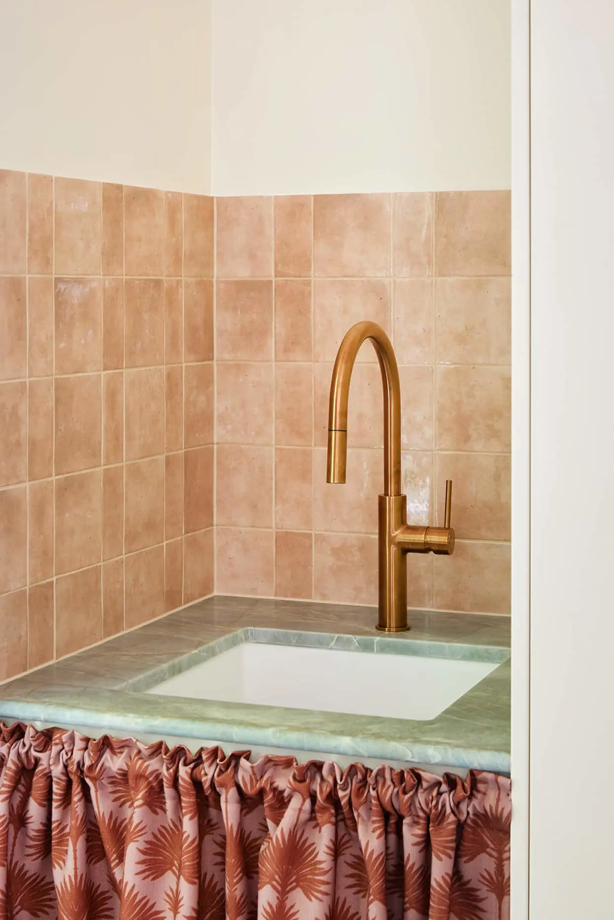 Modern bathroom sink with brass faucet, green marble countertop, terracotta tiles, and patterned curtain below.