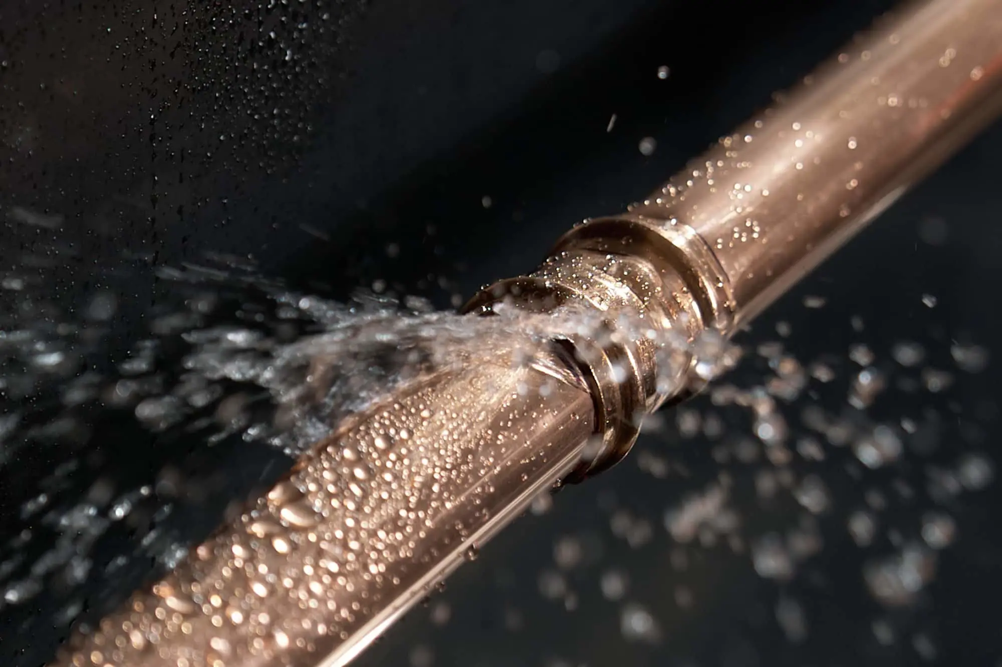 Water spraying from a broken copper pipe connection against a dark background, with droplets visible.