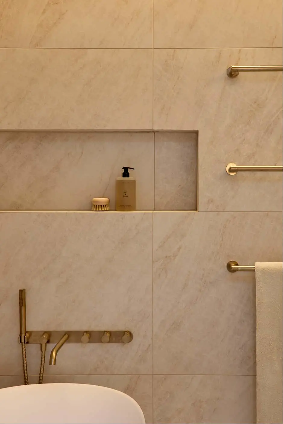 Bathroom wall with beige marble tiles, a recessed niche holding a soap dispenser and brush, brass towel rails, and wall-mounted faucet.