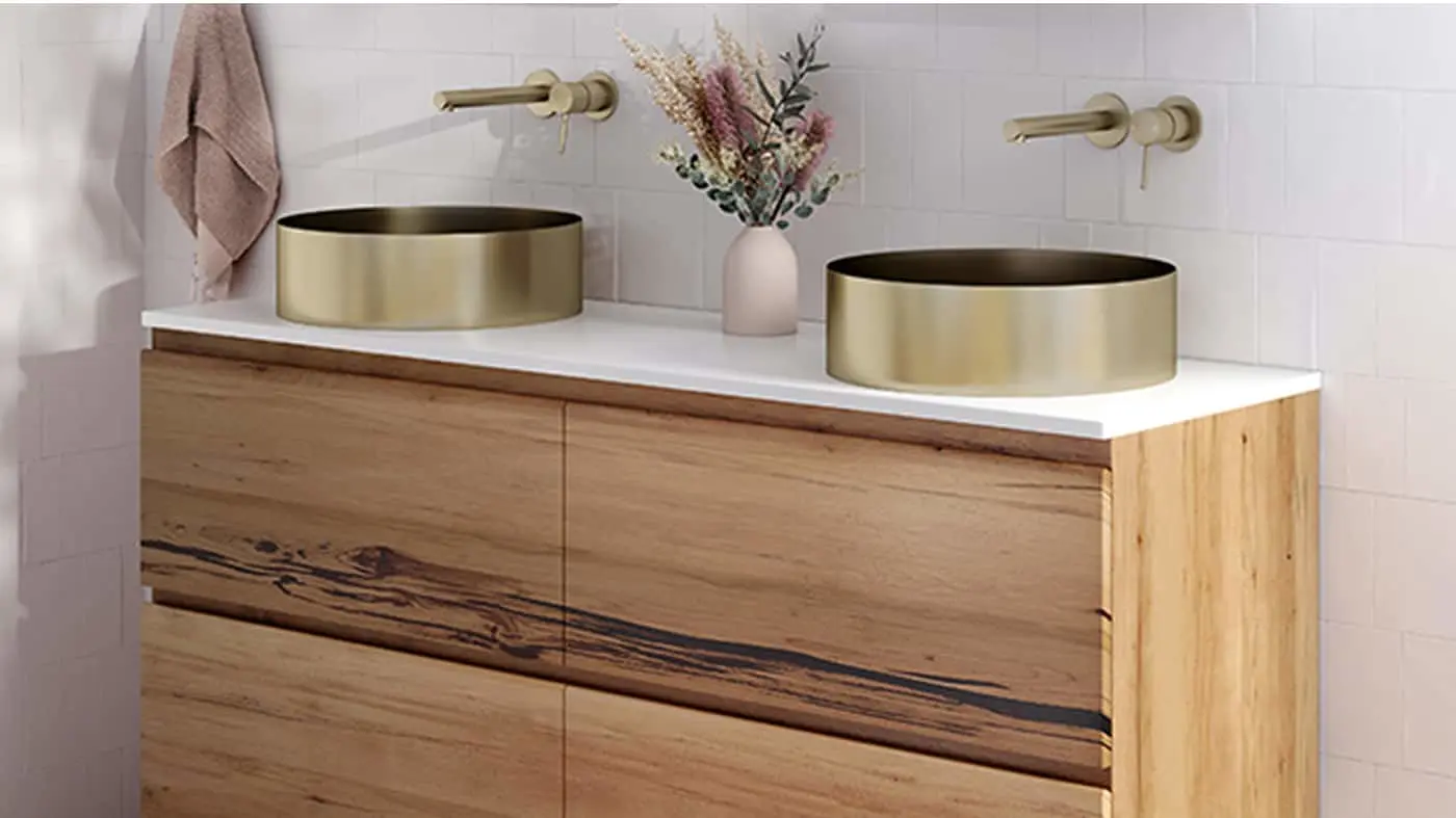 Modern bathroom vanity with wooden cabinet, white countertop, gold vessel sinks, wall-mounted faucets, and dried flowers.