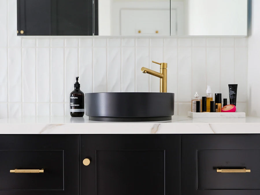 Modern bathroom with black vessel sink, gold faucet, white tile backsplash, and beauty products on marble countertop.