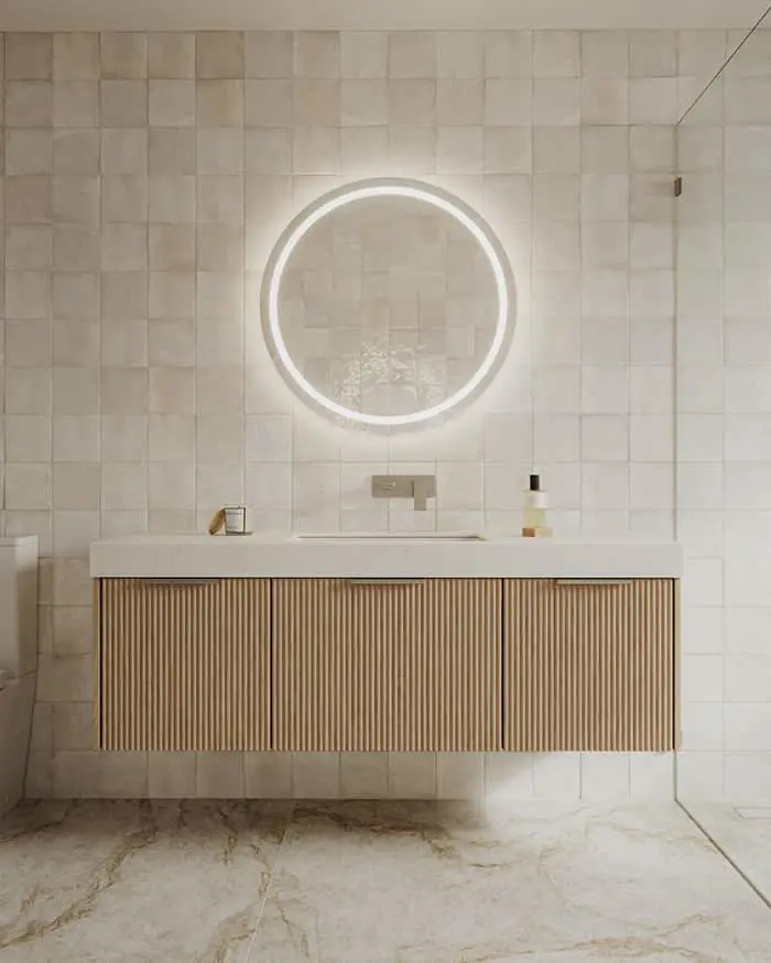 Modern bathroom with floating wooden vanity, white sink, illuminated round mirror, and cream tiled walls on marble floor.