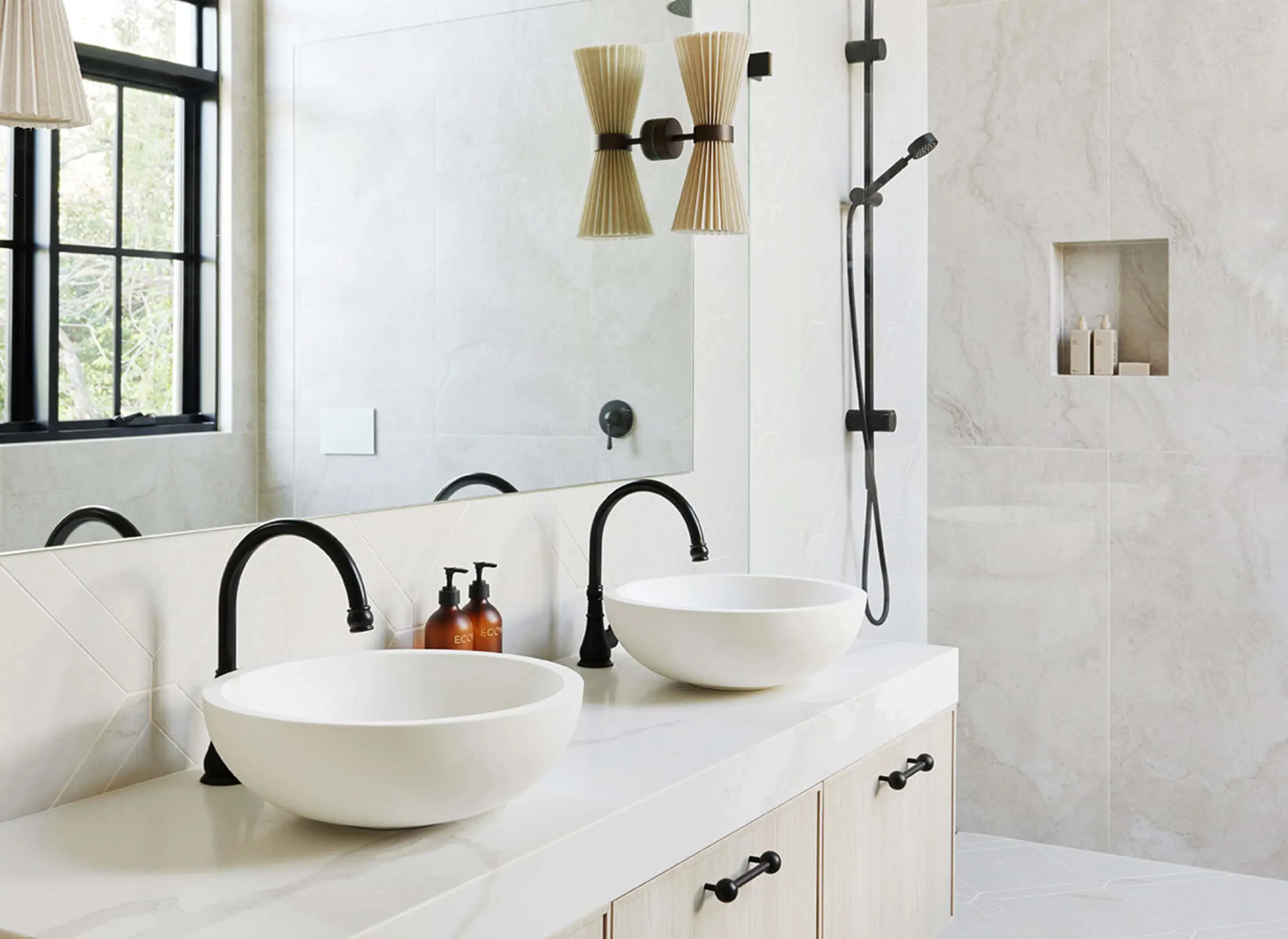 Modern bathroom with white vessel sinks, black fixtures, marble walls, and pleated wall sconces near a window.