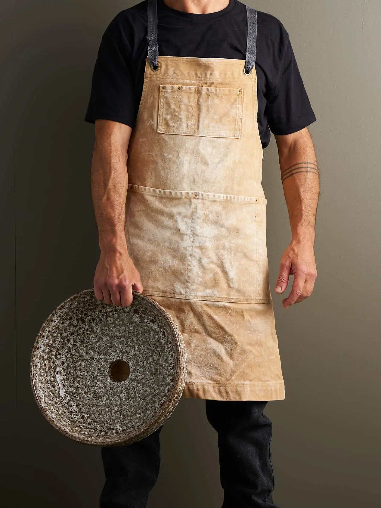 Person wearing a stained tan canvas apron with leather straps, holding a textured ceramic bowl with a hole in the center.
