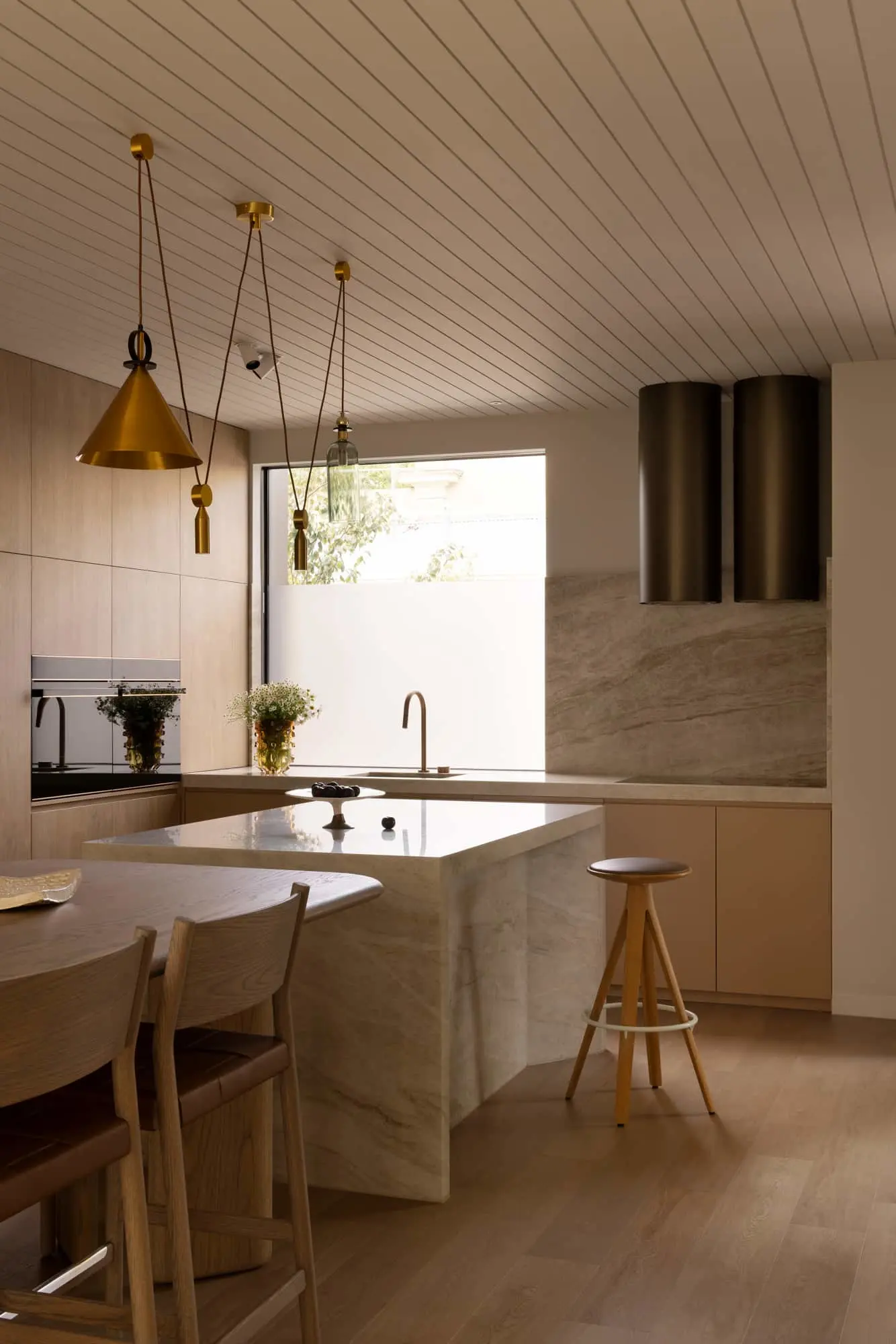 Modern kitchen with marble island, brass pendant lights, wooden stools, and large window providing natural light.