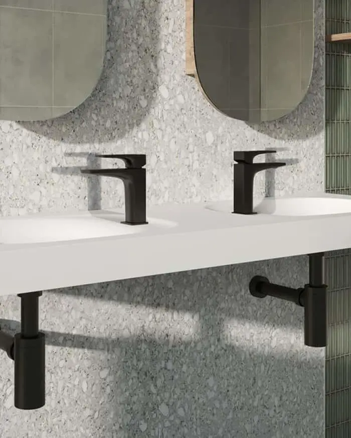 Modern bathroom vanity with two black faucets, white countertop, terrazzo wall, and oval mirrors.