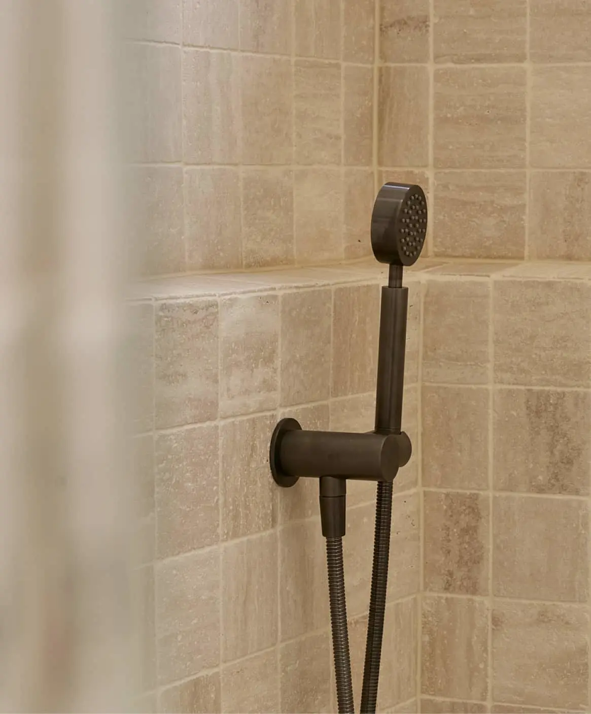 Gunmetal handheld shower head mounted on beige tiled shower wall with flexible metal hose.