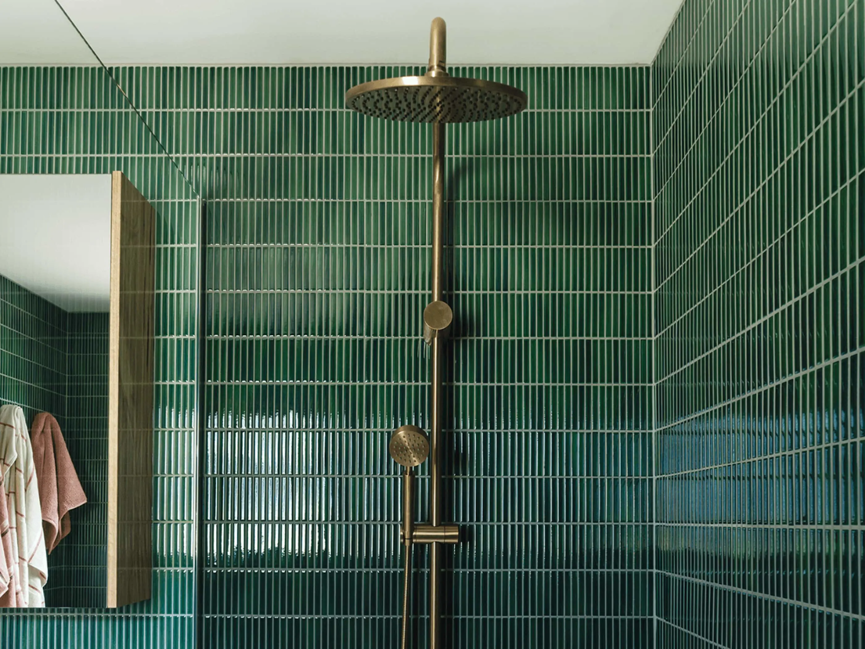 Modern shower with brass fixtures and green vertical tile walls, featuring a rainfall showerhead and handheld sprayer.