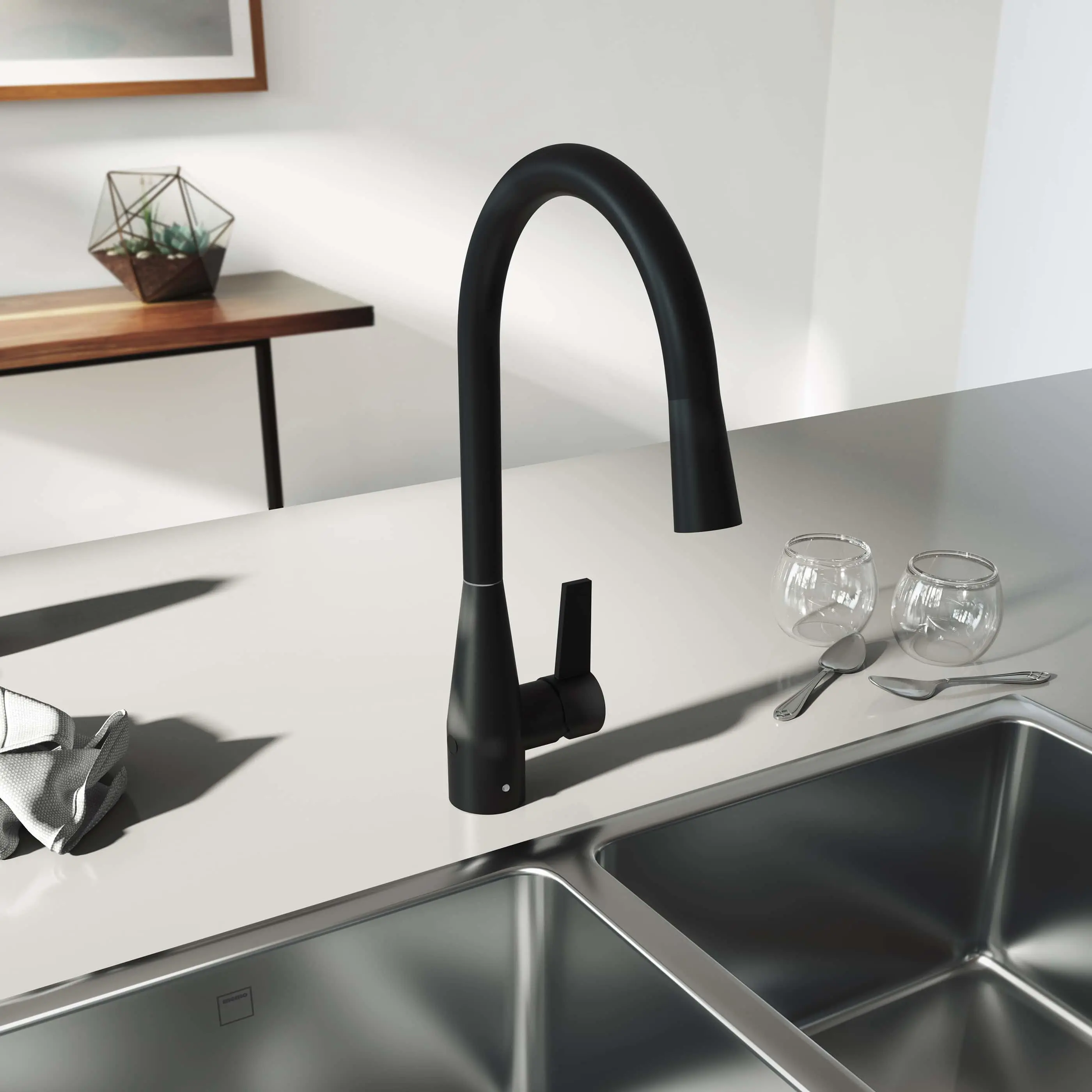 Modern black kitchen faucet with high arch over stainless steel sink, white countertop with glassware and small terrarium nearby.