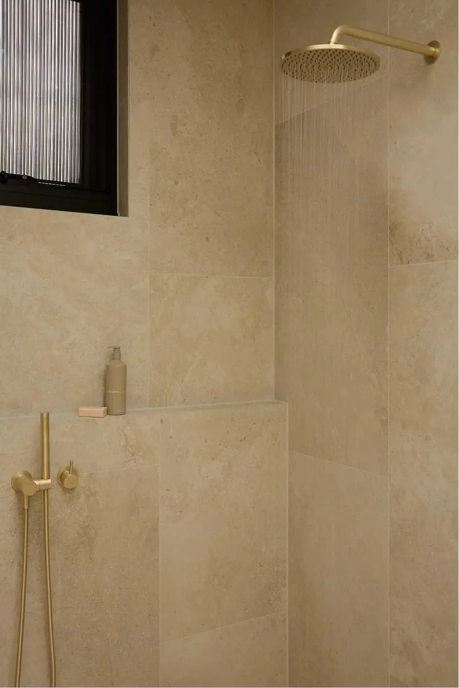 Beige tiled shower with brass rainfall showerhead and handheld fixture, built-in shelf with soap dispenser.