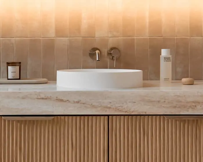 Modern bathroom with white vessel sink on marble countertop, beige tiled wall, and wooden cabinetry below.