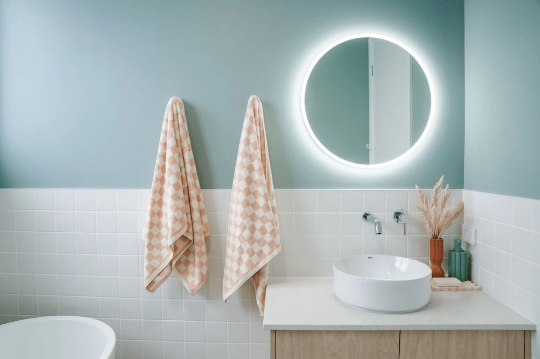 Modern bathroom with mint green walls, white tiles, backlit round mirror, peach checkered towels, and vessel sink.