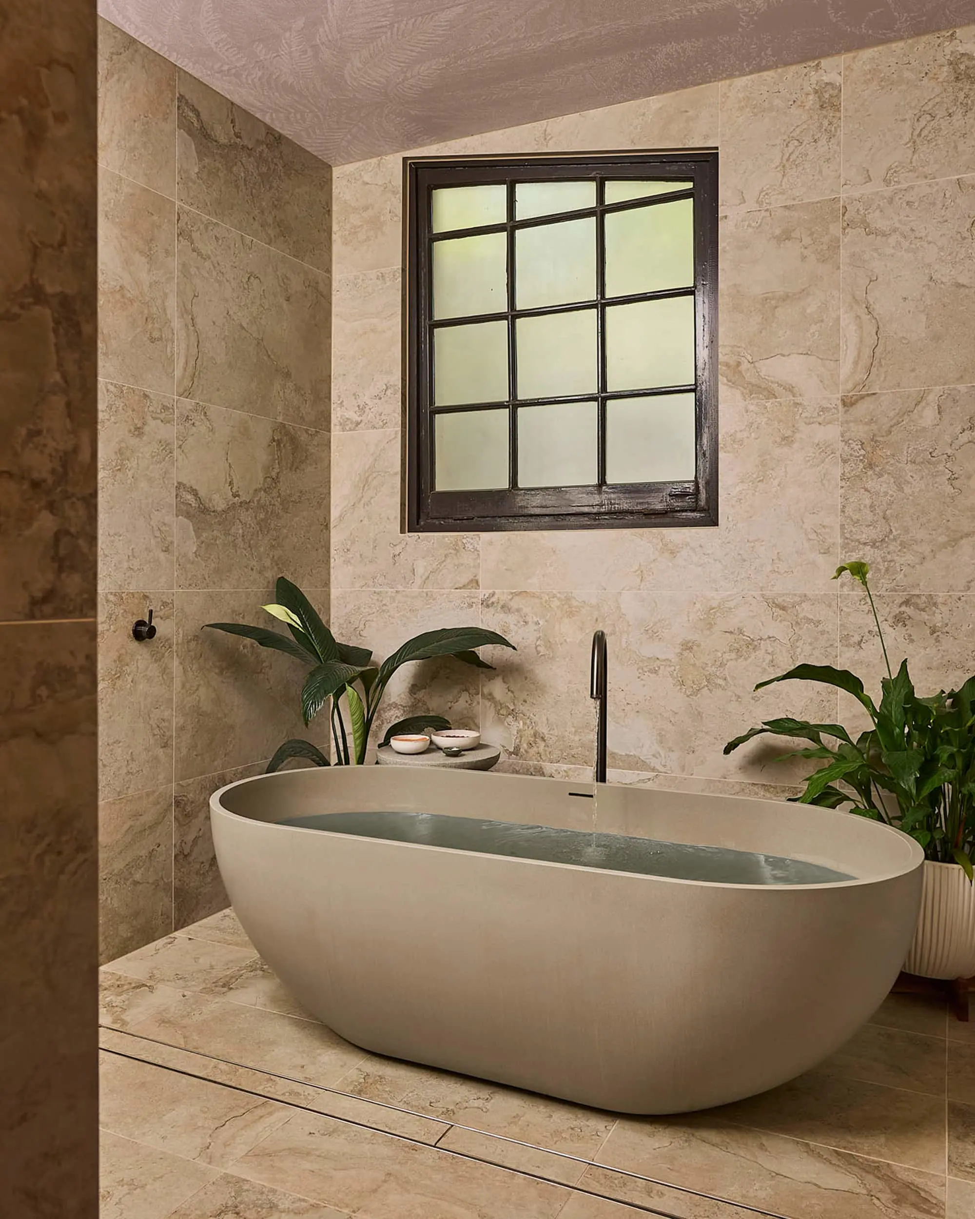 Modern bathroom with oval white bathtub, beige marble tiles, black-framed window, and green plants.