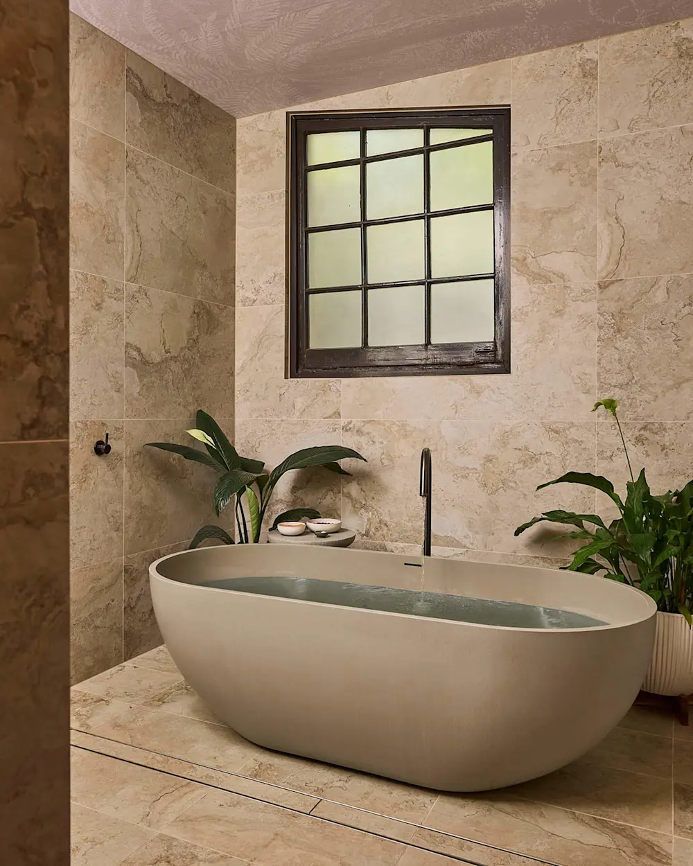 Modern bathroom with oval white bathtub, beige marble tiles, black-framed window, and green plants.