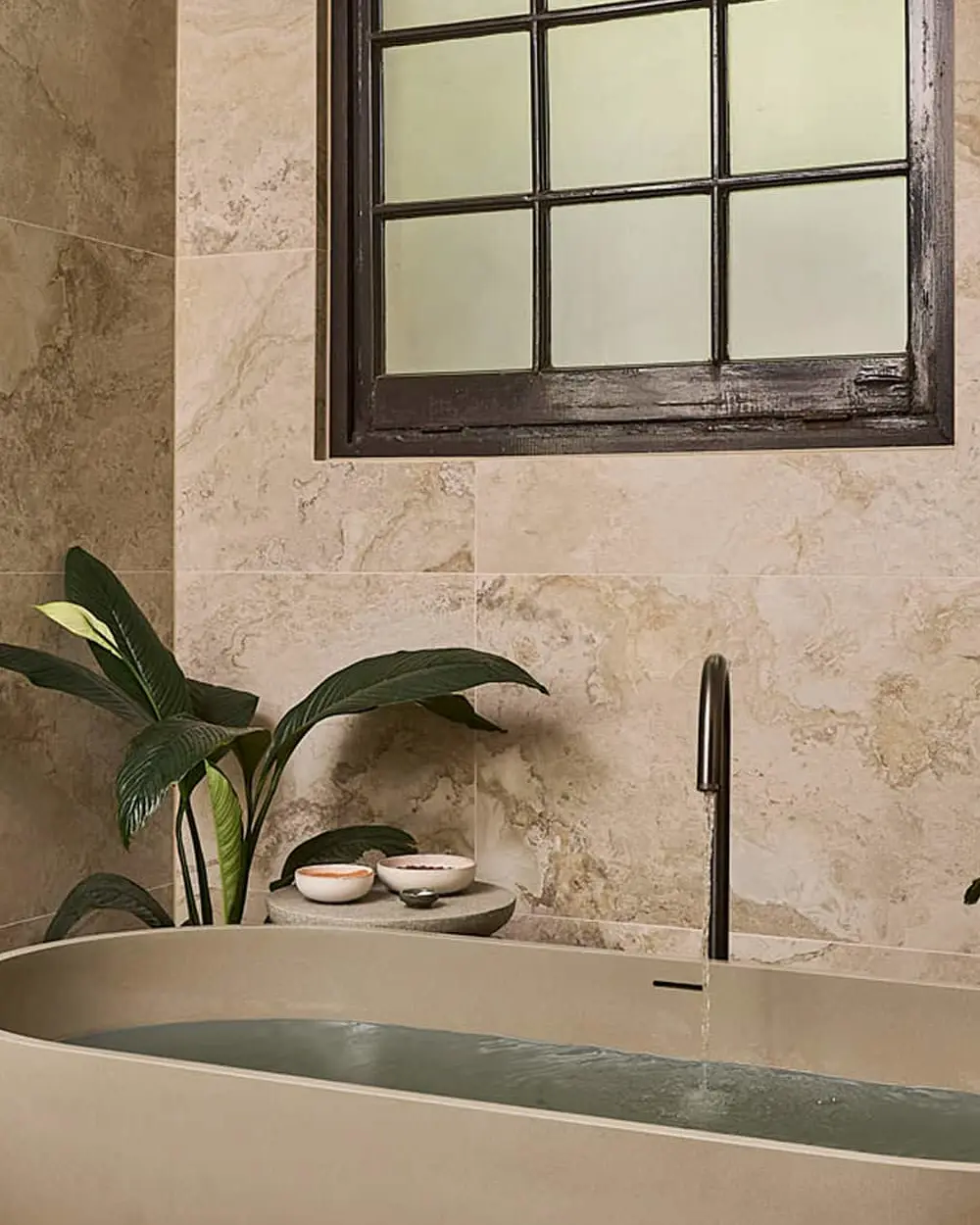 Modern bathroom with oval white bathtub, beige marble tiles, black-framed window, and green plants.