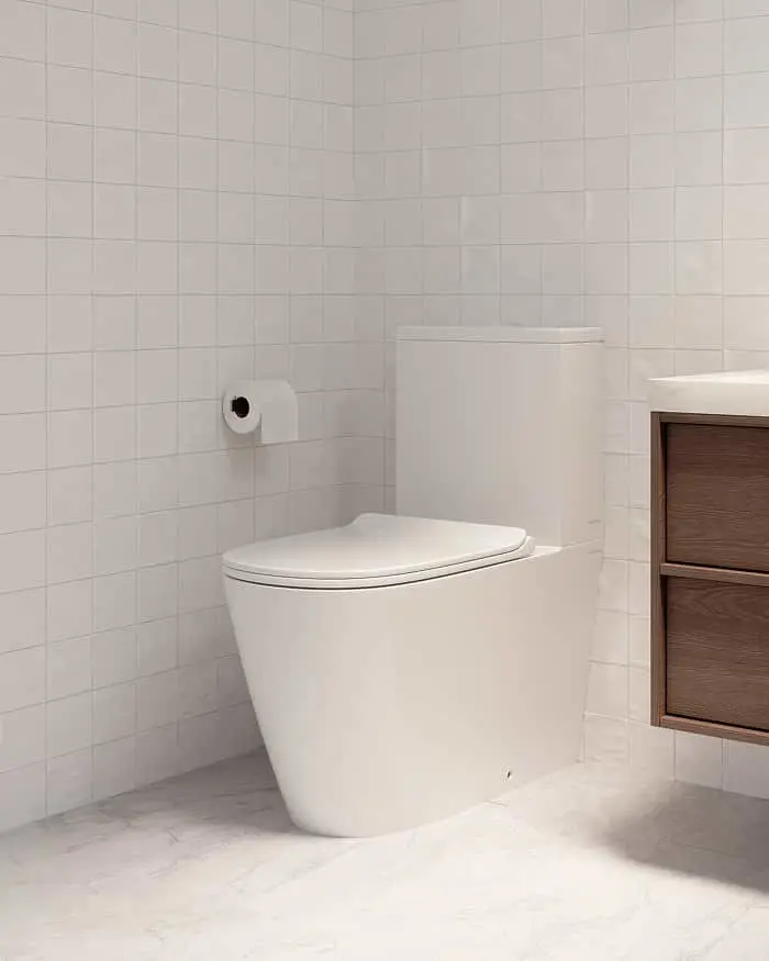 Modern white toilet in a minimalist bathroom with white tile walls and wooden vanity drawer visible.