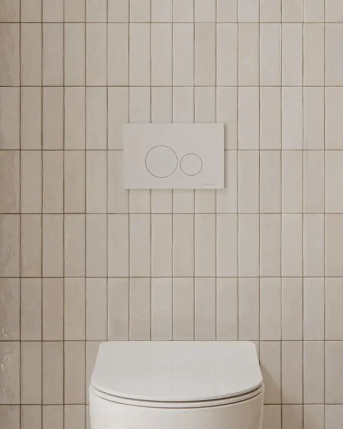 White wall-mounted toilet against beige tiled wall with a white dual-button flush plate mounted above.