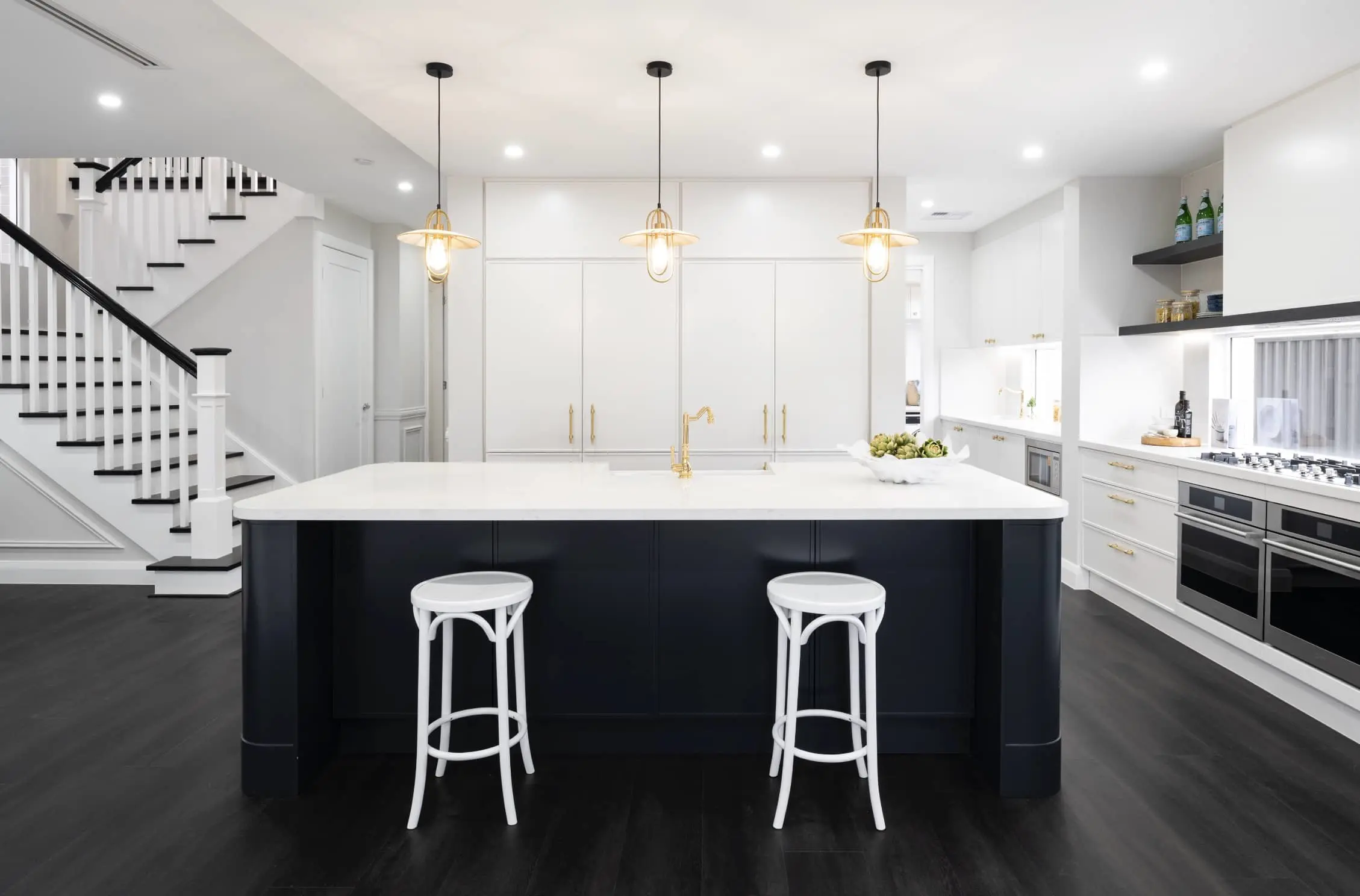 Modern white kitchen with navy island, white bar stools, pendant lights, and staircase visible to the left.