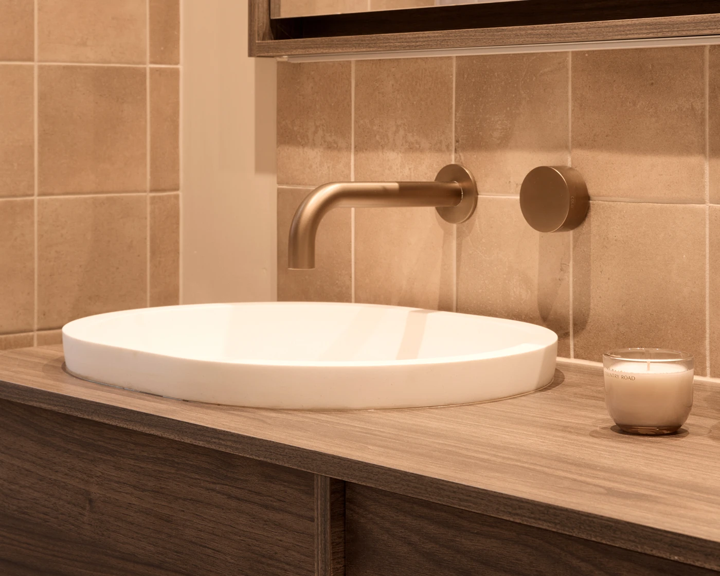 Modern bathroom sink with white oval basin, wall-mounted brass faucet, beige tiled wall, and small candle on wooden counter.