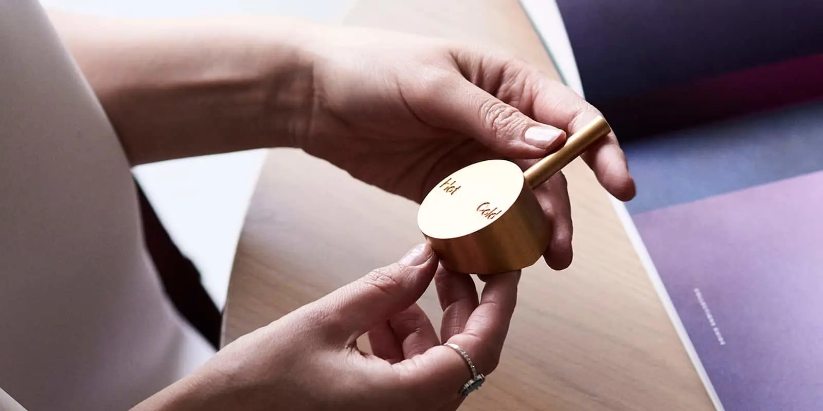 Two hands holding a small gold shower mixer object engraved with the words Hot and Cold on its top surface.