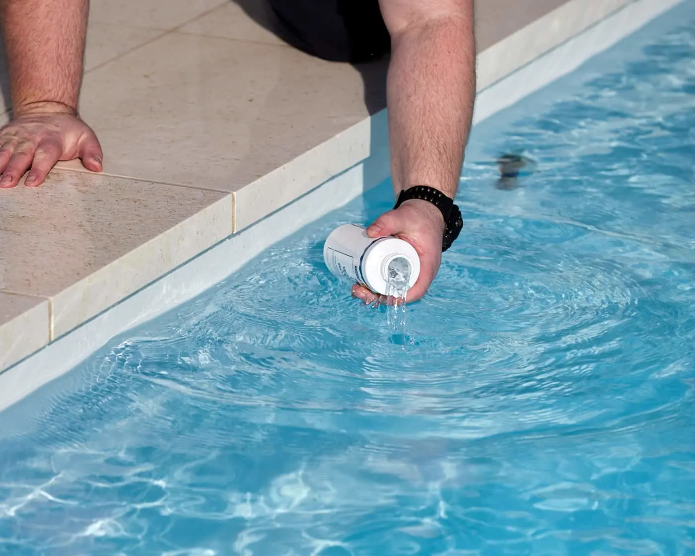 Person adding pool chemicals, pouring white container contents into blue swimming pool water from poolside edge.