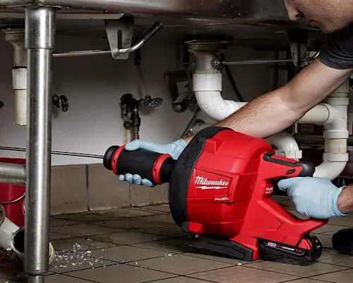 Plumber using red Milwaukee power drain cleaner on sink pipes under cabinet, wearing blue protective gloves.