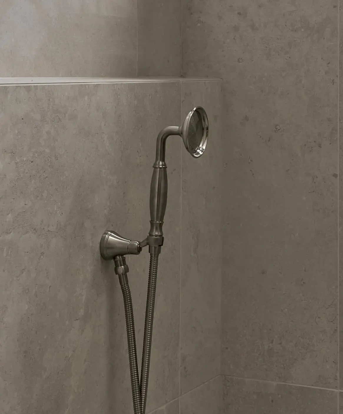 Brushed nickel handheld shower head mounted on gray stone tile wall with flexible metal hose.