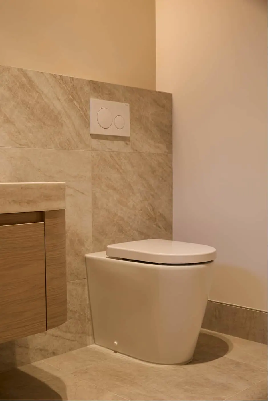 Modern bathroom with white toilet, beige marble tiles, and dual-button flush plate on wall.
