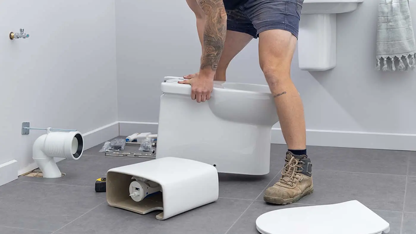 Person installing a toilet in bathroom with plumbing parts visible on gray tile floor.