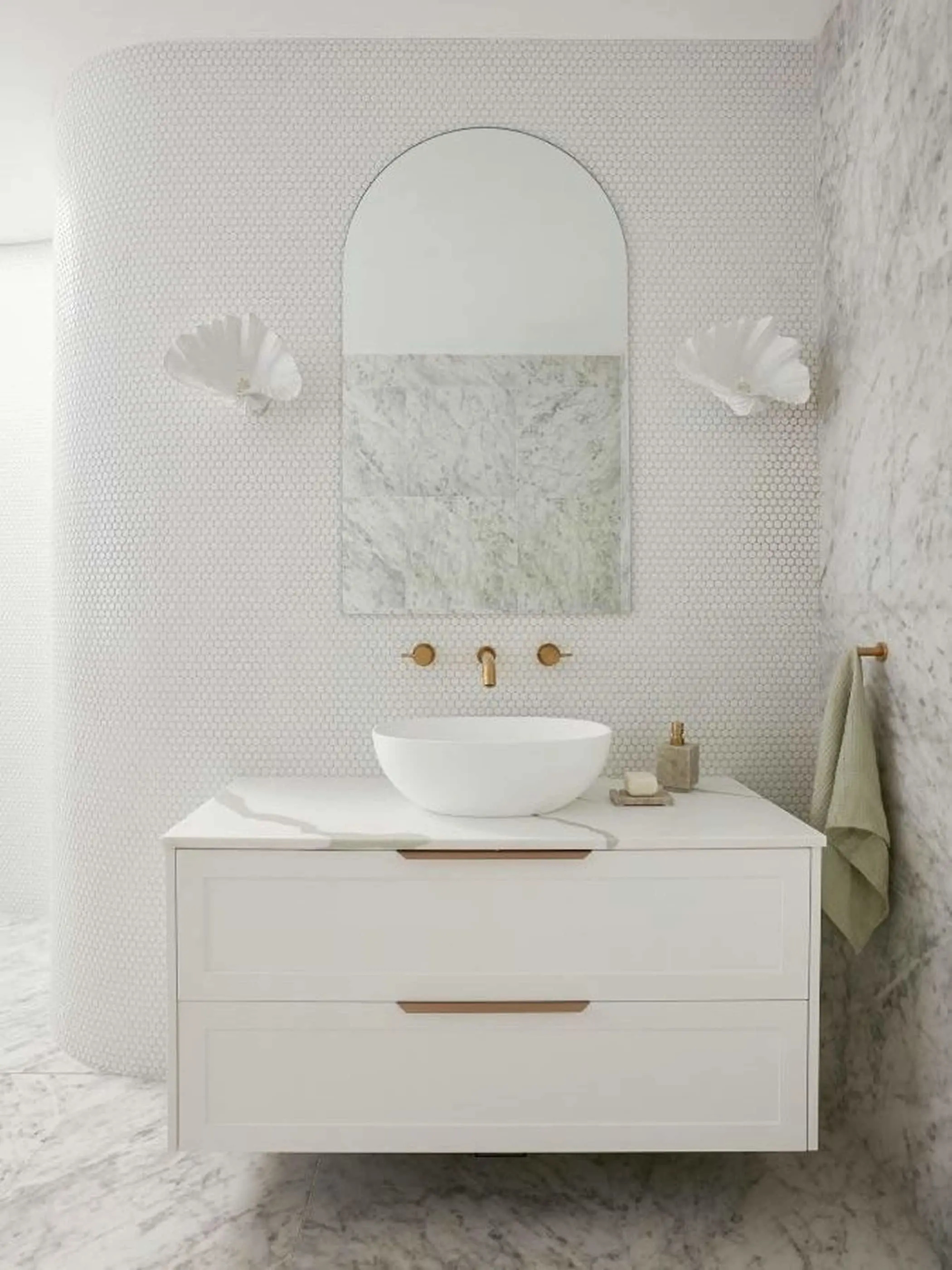 Elegant white bathroom with marble accents, arched mirror, vessel sink, brass fixtures, and shell-shaped sconces.