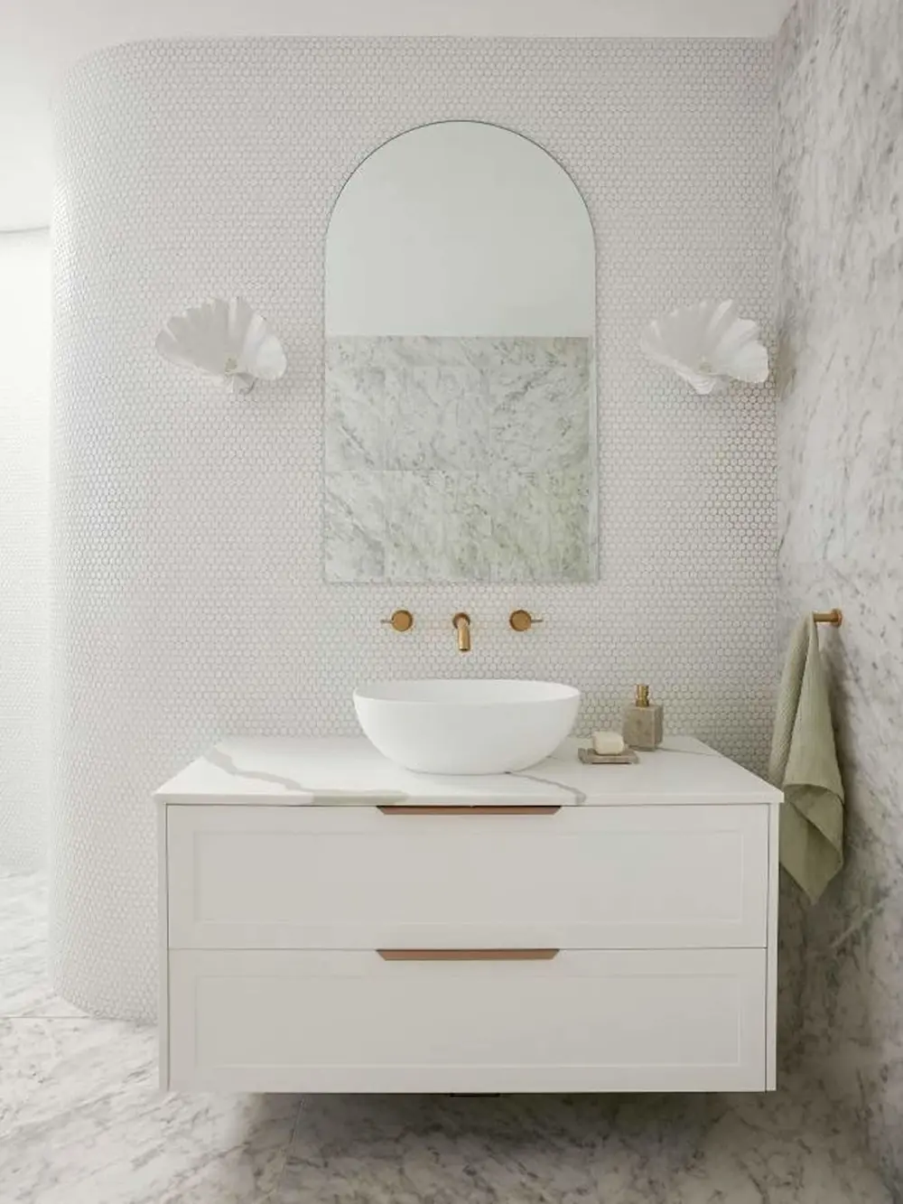 Elegant white bathroom with marble accents, arched mirror, vessel sink, brass fixtures, and shell-shaped sconces.