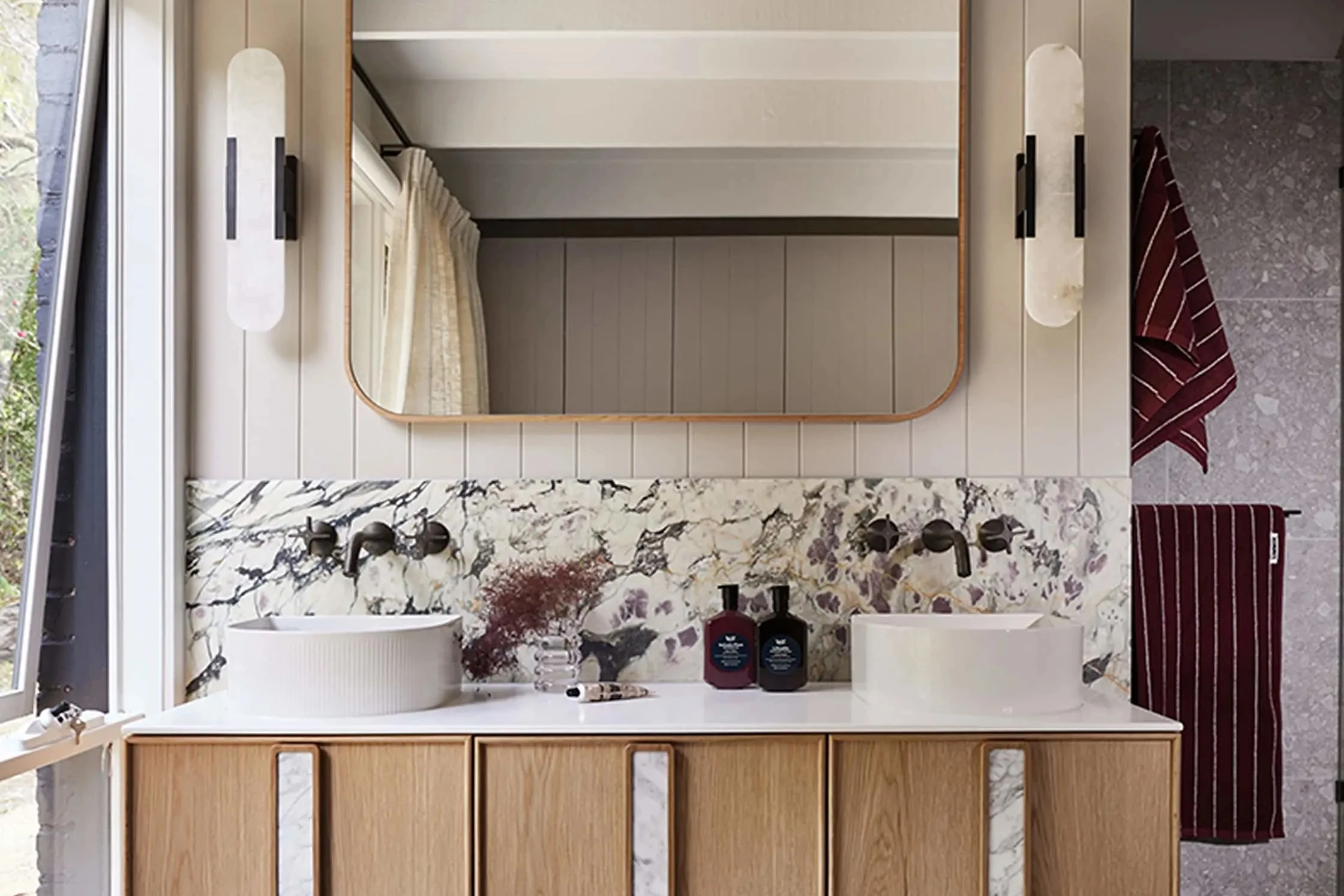 Modern bathroom with wooden floating vanity, marble backsplash, round vessel sinks, and gold-framed mirror with wall sconces.