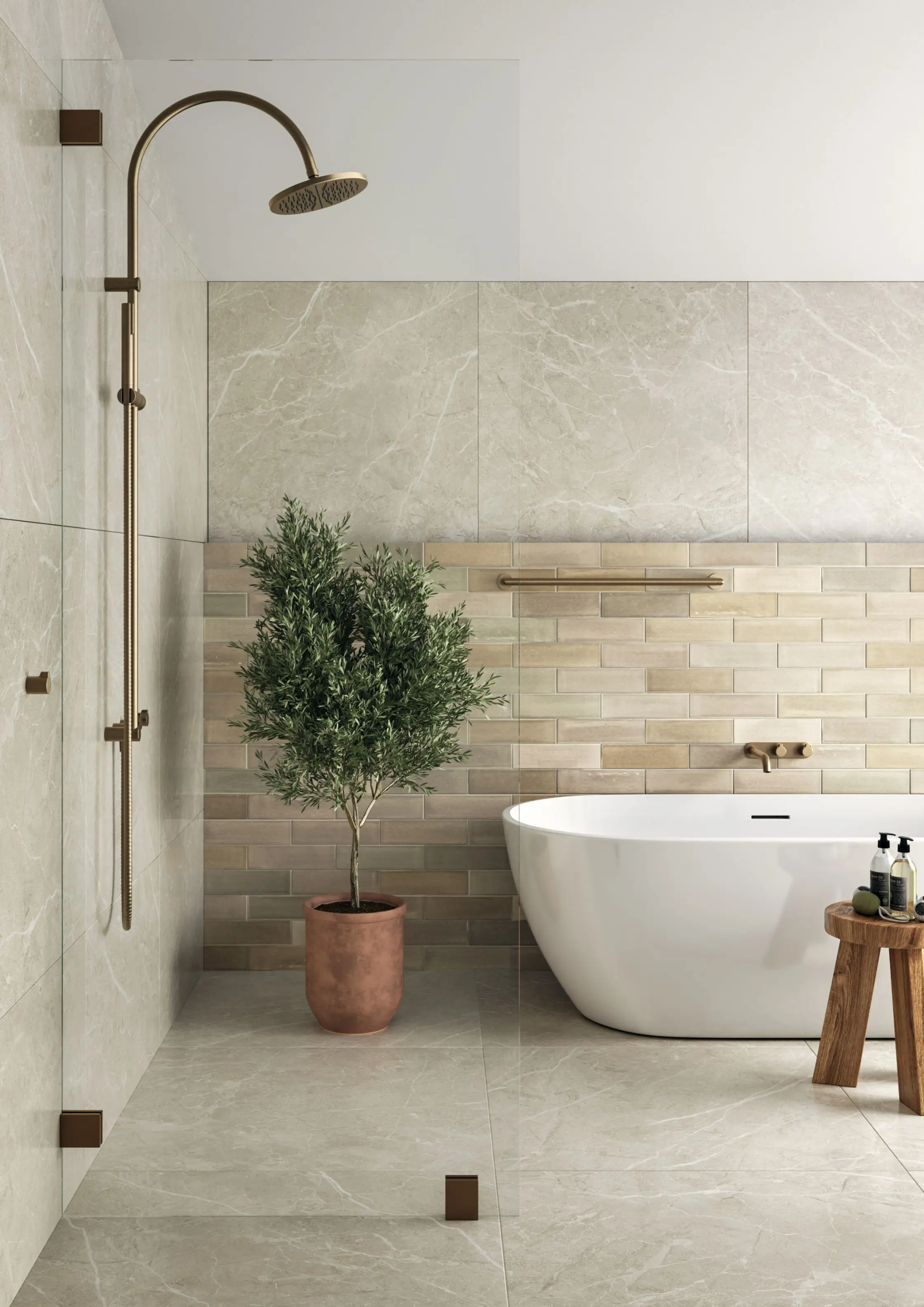 Bathroom with beige marble tiles, a white freestanding bathtub, brass shower fixtures, a potted green tree, and a wooden stool.