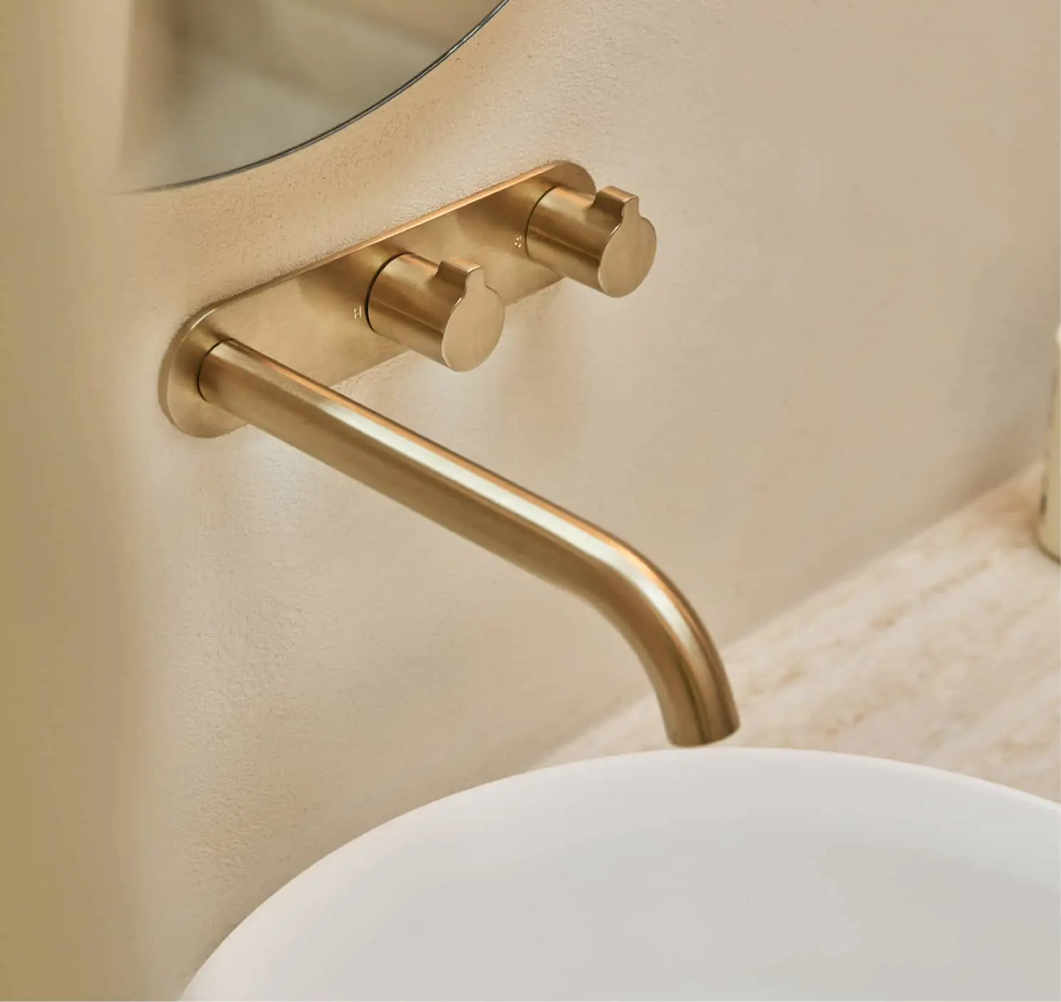 Brushed gold wall-mounted basin tap with two cylindrical handles above a white round sink on a beige wall.