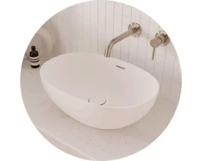 Oval white bathroom sink on light countertop with wall-mounted brushed metal faucet against white paneled wall.