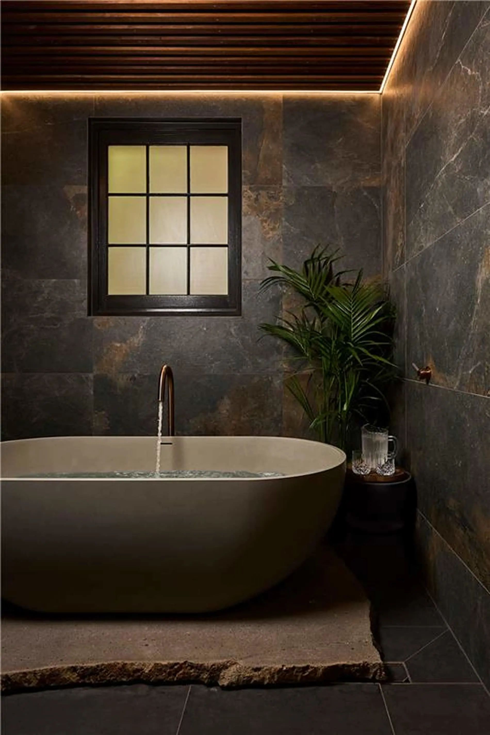 Modern bathroom with oval white tub, dark stone walls, wooden ceiling, window, and potted palm plant.