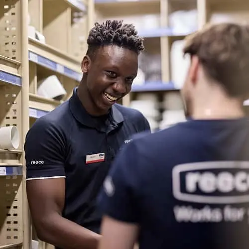 Two employees in navy blue polo shirts with company logos conversing in a storage area with shelving units