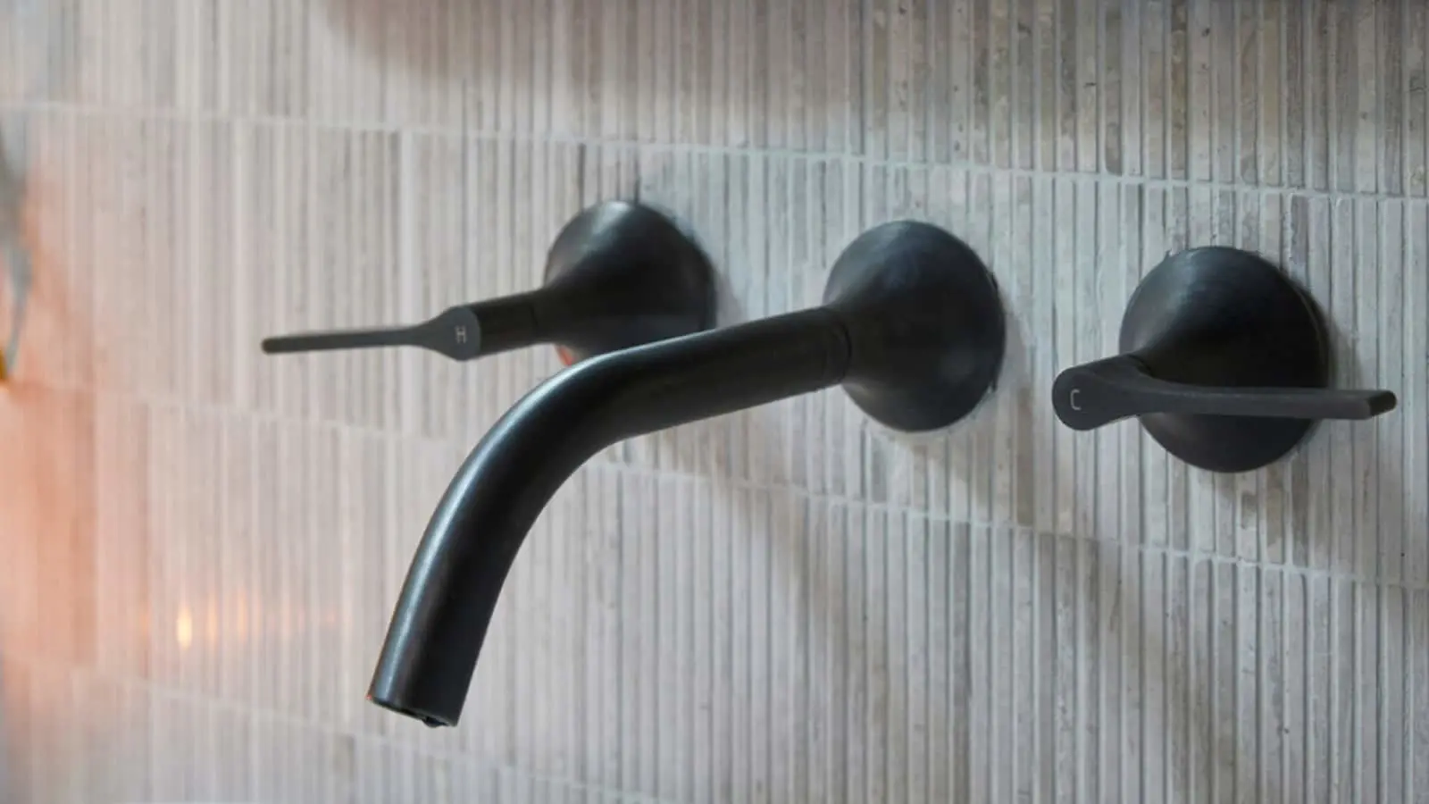 Modern black wall-mounted bathroom faucet with three controls against textured light gray tile wall.