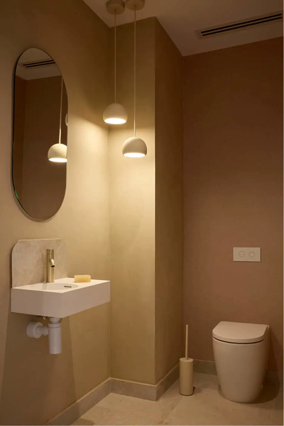 Small bathroom with warm beige walls, wall-mounted sink, oval mirror, pendant lights, and a wall-hung toilet.