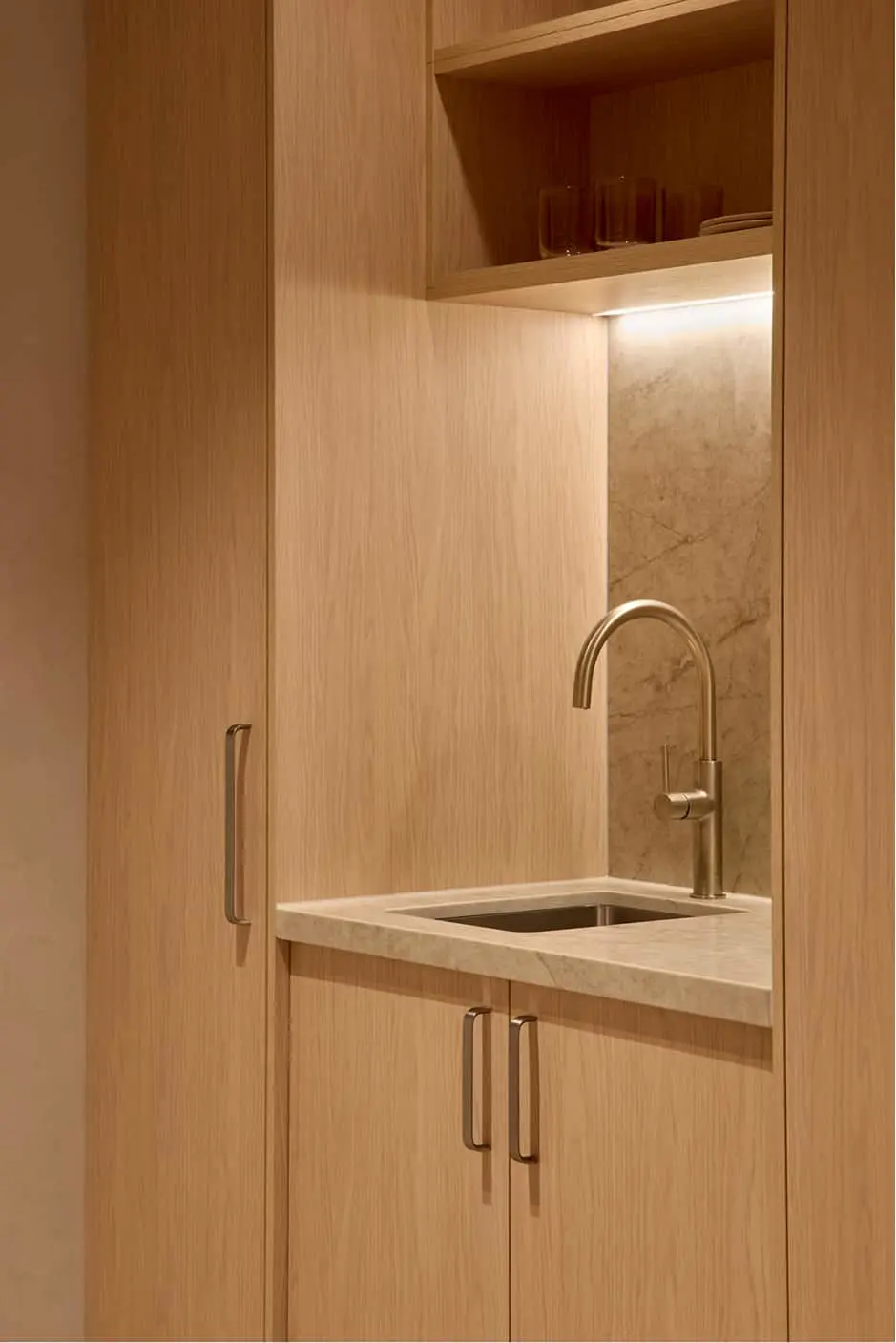 Light wood cabinetry with sink, faucet, and open shelf holding glassware above a marble backsplash.