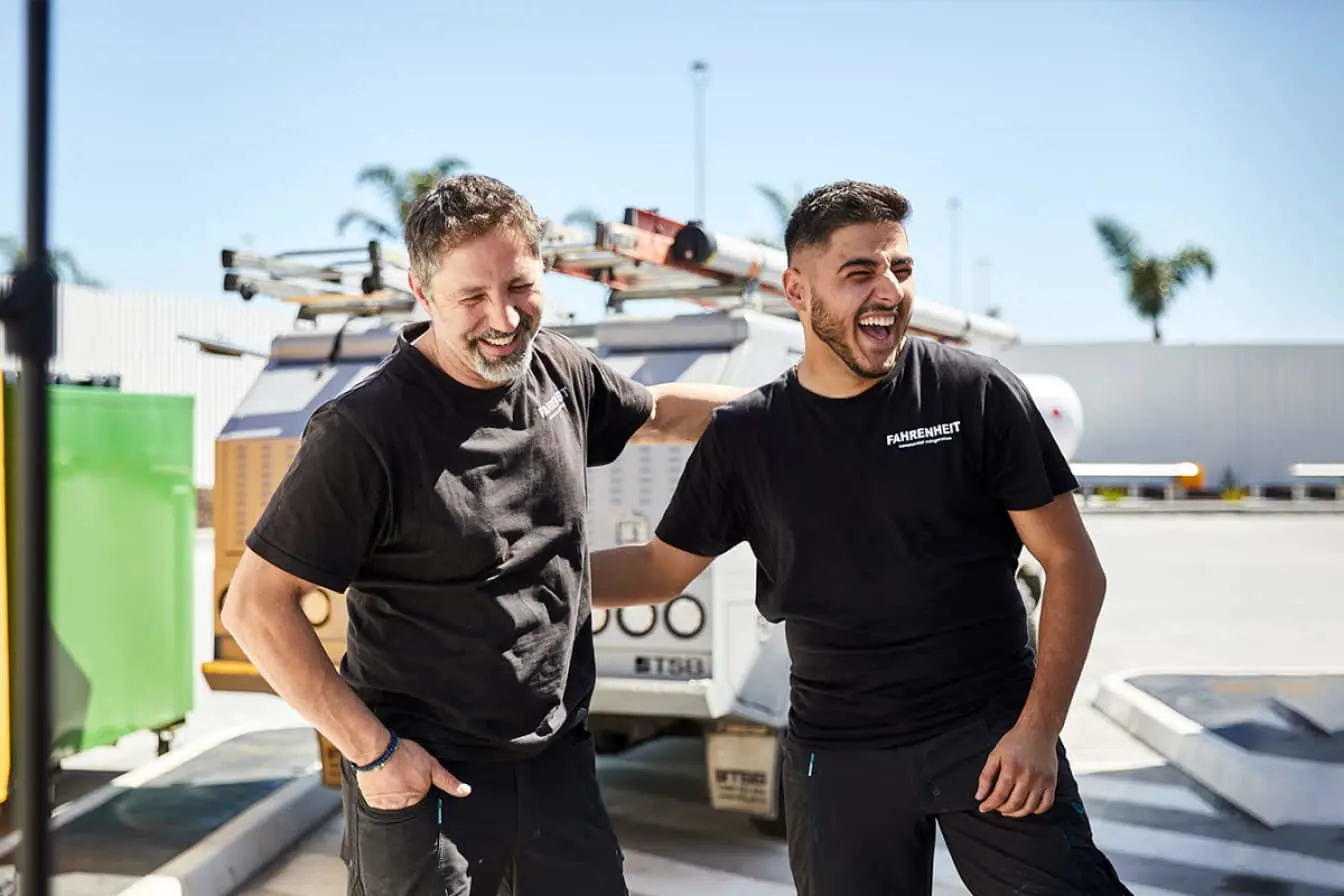 Two workers in black t-shirts laughing together outdoors, with a service vehicle and equipment visible in background.