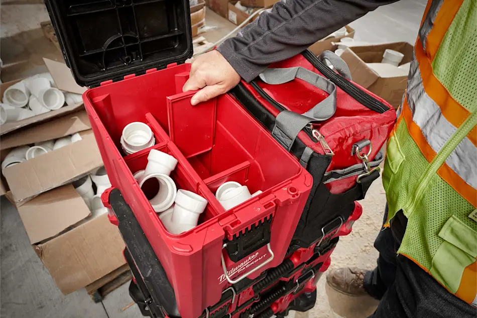 Person opening red Milwaukee toolbox containing white plumbing fittings, with tool bag and cardboard boxes nearby.